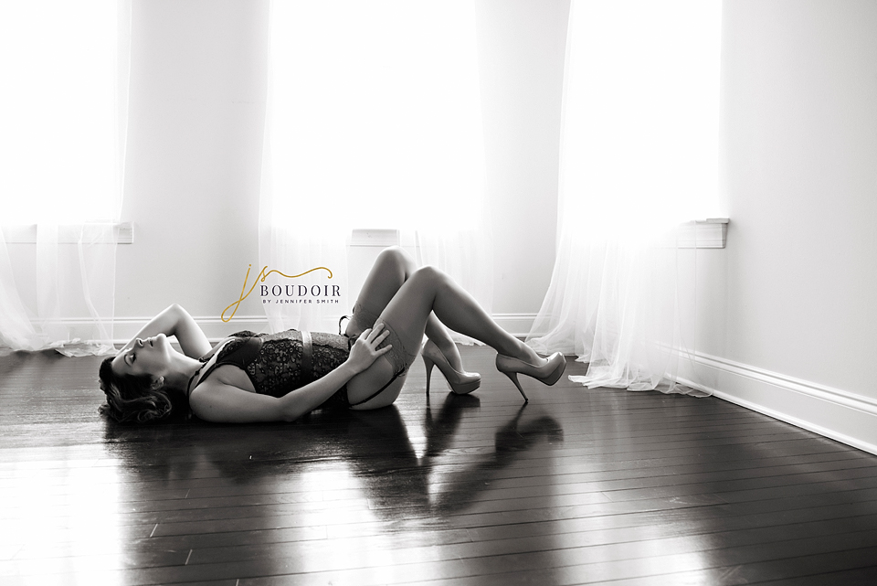 A woman in lingerie and heels lies on her back on a wooden floor, bathed in soft light from two sheer-curtained windows, conveying elegance and serenity.